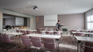 A refined space with grey carpet, white walls, and natural light cast from the left-side windows. Set up in classroom style, a ceiling-mounted projector beams towards a large screen on the top wall's center. On the left is a breakfast bar with food and refreshments. A great choice for event spaces in Dublin.