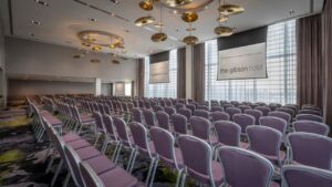 A spacious room for grand events featuring numerous rows of purple chairs arranged in theatre style, facing two expansive projector screens. Behind the screens are several large windows from which natural light pours, adorned with light brown curtains. Gold lighting fixtures hang from the ceiling to make this one of the most luxurious venues in Dublin, Ireland.