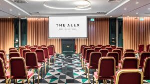Stunning room with a green and white checkered carpet. The stylish brown divides the space, leading to a large screen suspended from the roof in front of rows of comfortable red and gold chairs, arranged theatre-style. A fantastic choice for conference venues in Dublin