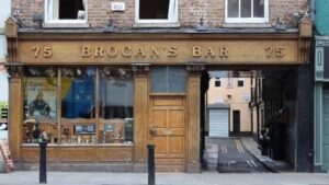 The exterior of Brogans Bar presents a charming wooden facade, evoking a timeless appeal. To the left, three rectangular windows showcase a curated collection of old brewery memorabilia, offering a glimpse into the establishment's rich history. A wooden door, exuding a classic allure, serves as the entrance. Alongside, a lengthy alleyway runs to the right, all beneath the welcoming presence of the pub's signage. This harmonious composition invites patrons to step into a place where tradition and character converge in Temple Bar Dublin.
