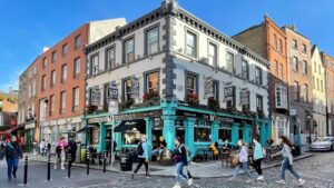 The Norseman's exterior, nestled on a street corner, boasts cobblestone paths on both sides. Outside, a blend of tables, barrels, chairs, and stools create a lively setting. The scene is bustling with people strolling by, capturing the essence of a vibrant and animated atmosphere in Temple Bar Dublin.