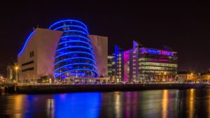 Nighttime view of the Dublin Convention Centre. The building's architecture creates a striking visual, with a luminous ring encircling the windows. Positioned alongside the quay, its reflection glistens on the water, creating a stunning scene.